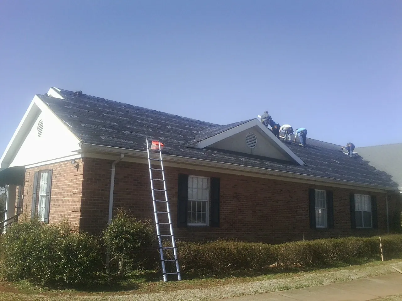 Crew tearing shingles off a brick ranch roof with a ladder set — Emergency Roof Repair work by our crew in Swartz Creek