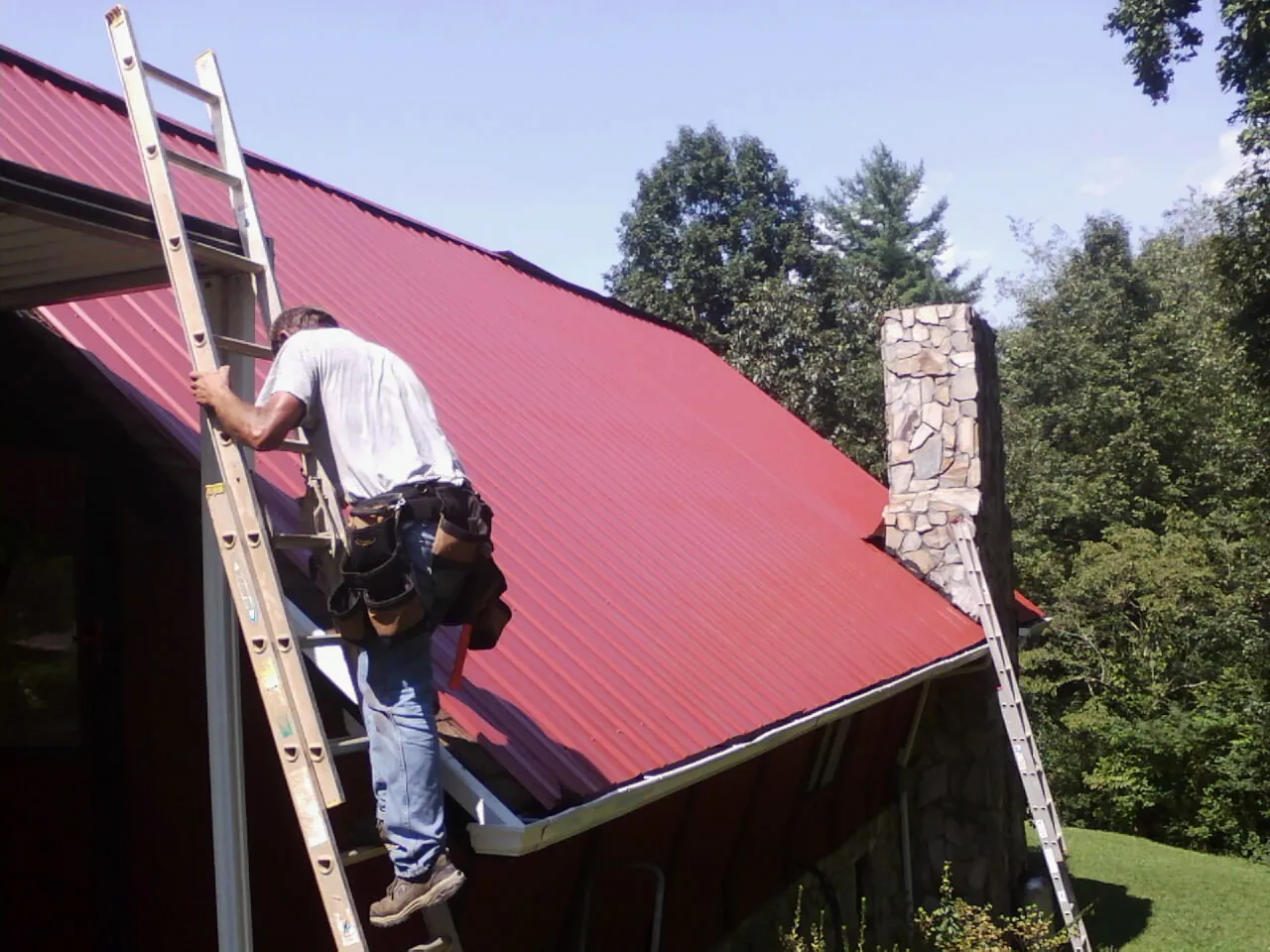 Roofer on a ladder beside a red metal roof and stone chimney — Soffit & Fascia Repair by Roofing Brothers in Swartz Creek